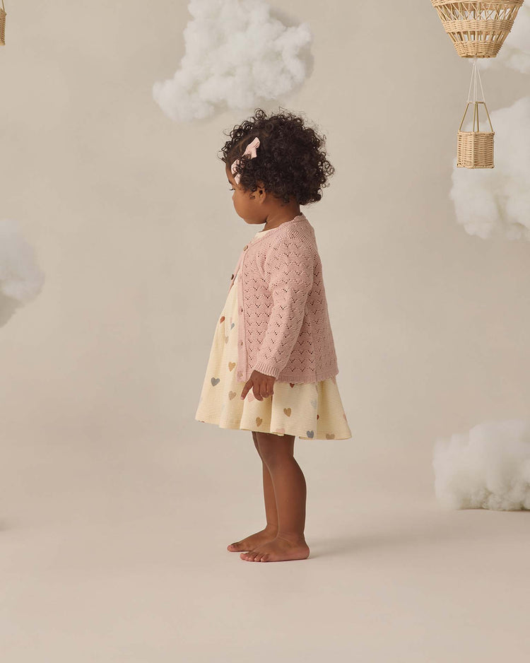 A young child with curly hair stands in profile, wearing a pink cardigan and the quincy mae darla dress in confetti hearts. Fluffy clouds and wicker baskets hang above, creating a whimsical scene.