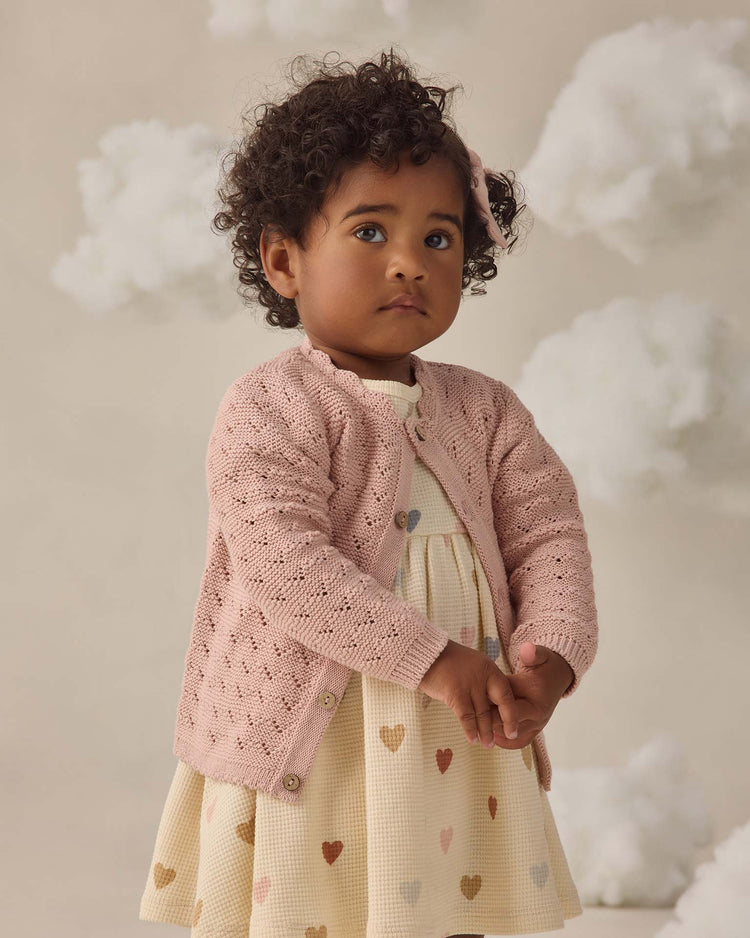 A young child with curly hair wears the quincy mae scalloped cardigan in blush over a cream dress with multicolored hearts. She stands against a light background with fluffy cloud decorations, gazing slightly to the side.