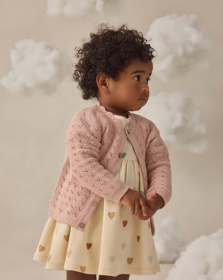 A young child with curly hair wears the quincy mae scalloped cardigan in blush over a cream dress with multicolored hearts, standing against a backdrop of fluffy white clouds.
