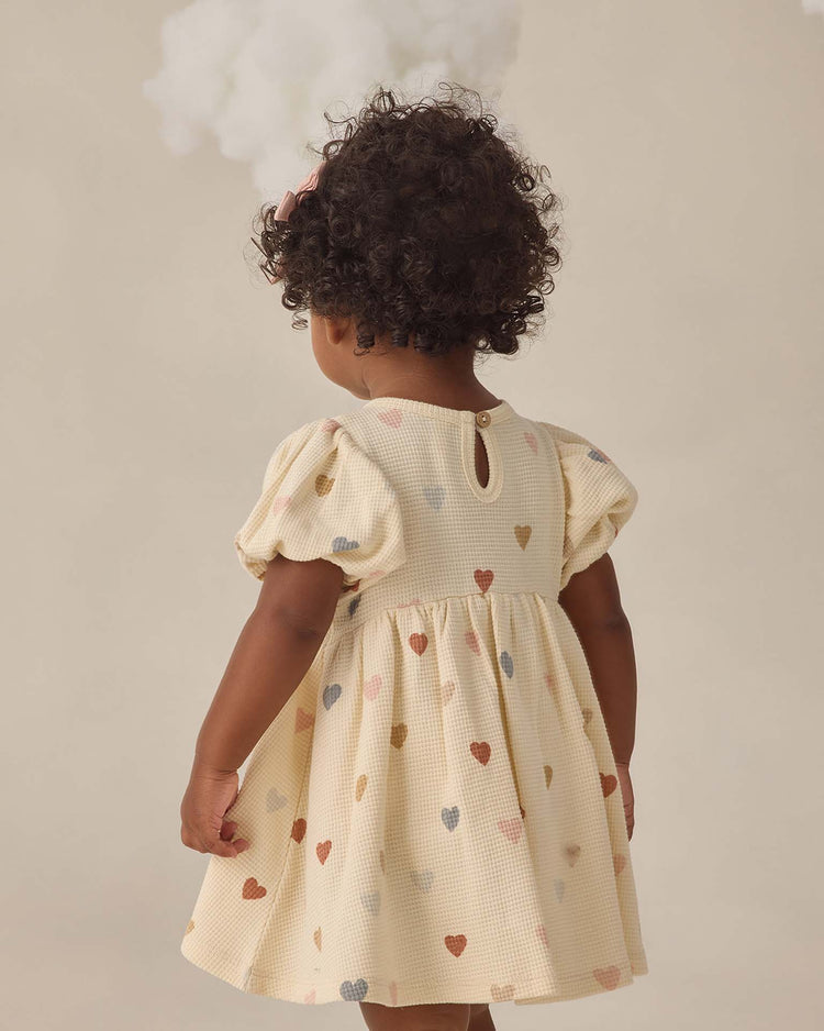 A young child with curly hair, seen from behind, wears the quincy mae darla dress in confetti hearts with puffed sleeves. A white fluffy cloud hovers above against a neutral background.