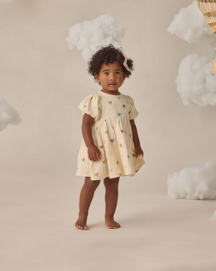 A young child with curly hair wears the Quincy Mae Darla Dress in Confetti Hearts, made from cream organic cotton with colorful heart motifs, standing barefoot against a beige background with fluffy white clouds.