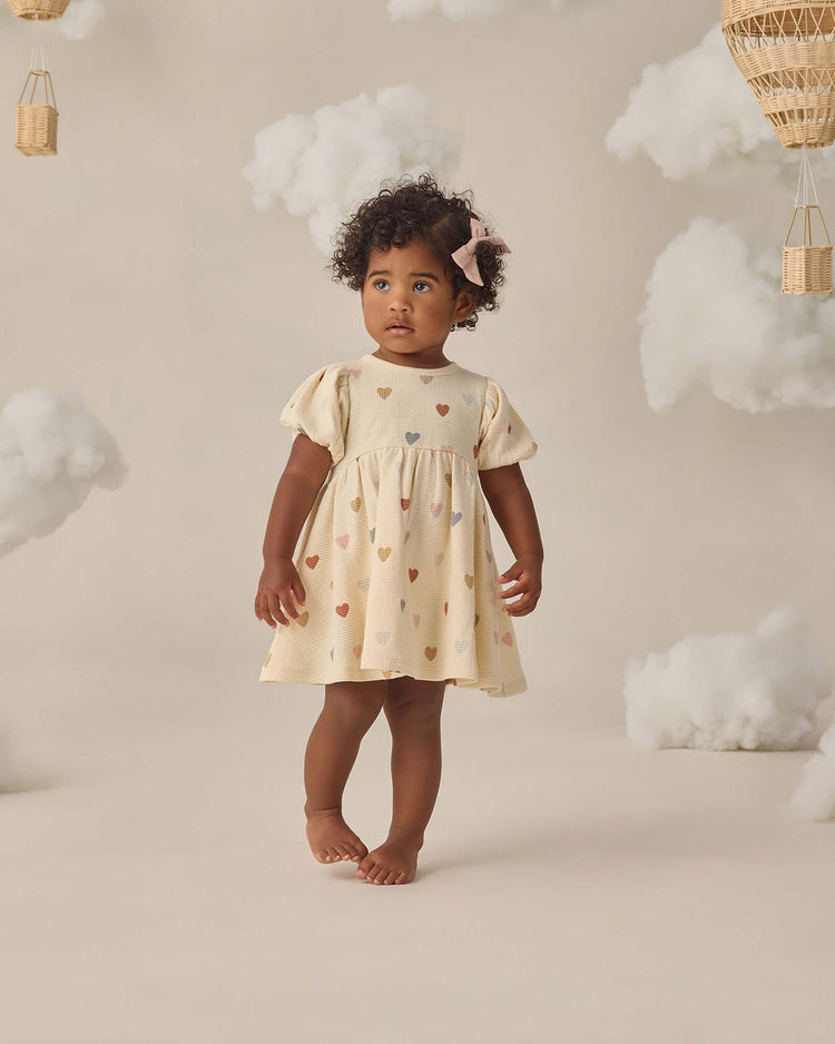 A young child with curly hair stands barefoot on a soft beige floor, wearing the quincy mae darla dress in confetti hearts, surrounded by fluffy cloud props and wicker baskets decorated with colorful hearts.