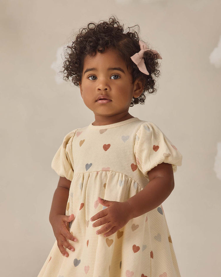 A young child with curly hair and a pink bow wears the quincy mae darla dress in confetti hearts, crafted from organic cotton, standing against a soft neutral background.