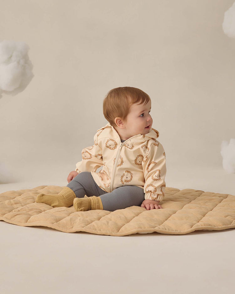A baby sits on a beige quilted mat, wearing a cream hooded lion print jacket, cozy yellow socks, and Quincy Mae’s ribbed legging in ocean. The light background features soft, cloud-like shapes.