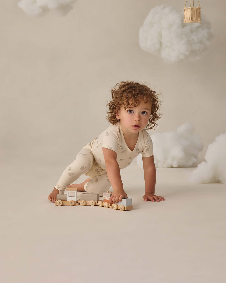 A toddler with curly hair wears the quincy mae bamboo short sleeve pajama set in elephants while playing on the floor with a wooden toy train, surrounded by white fluffy clouds for a dreamy atmosphere.