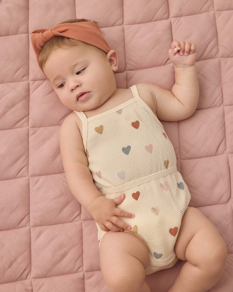 A baby in the quincy mae waffle cinch romper in confetti hearts and a rust-colored headband lies on a quilted pink blanket, gazing to the side with one arm raised.
