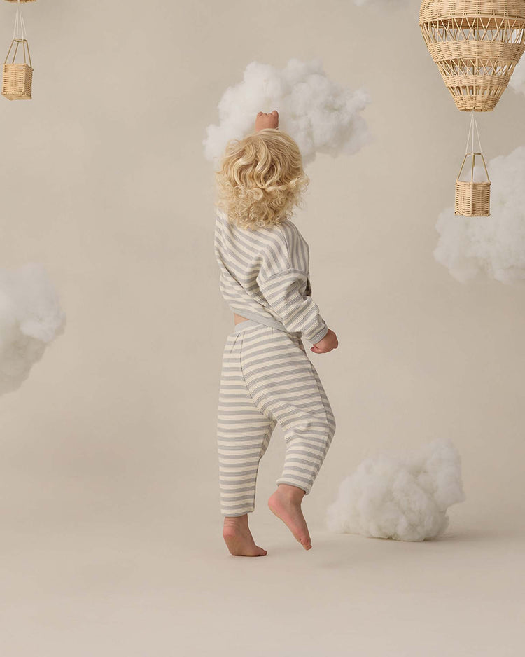 A small child with curly blonde hair wears the quincy mae waffle sweater set in sky stripe, standing barefoot and reaching toward a fluffy white cloud in a whimsical setting with hanging wicker baskets and soft clouds.