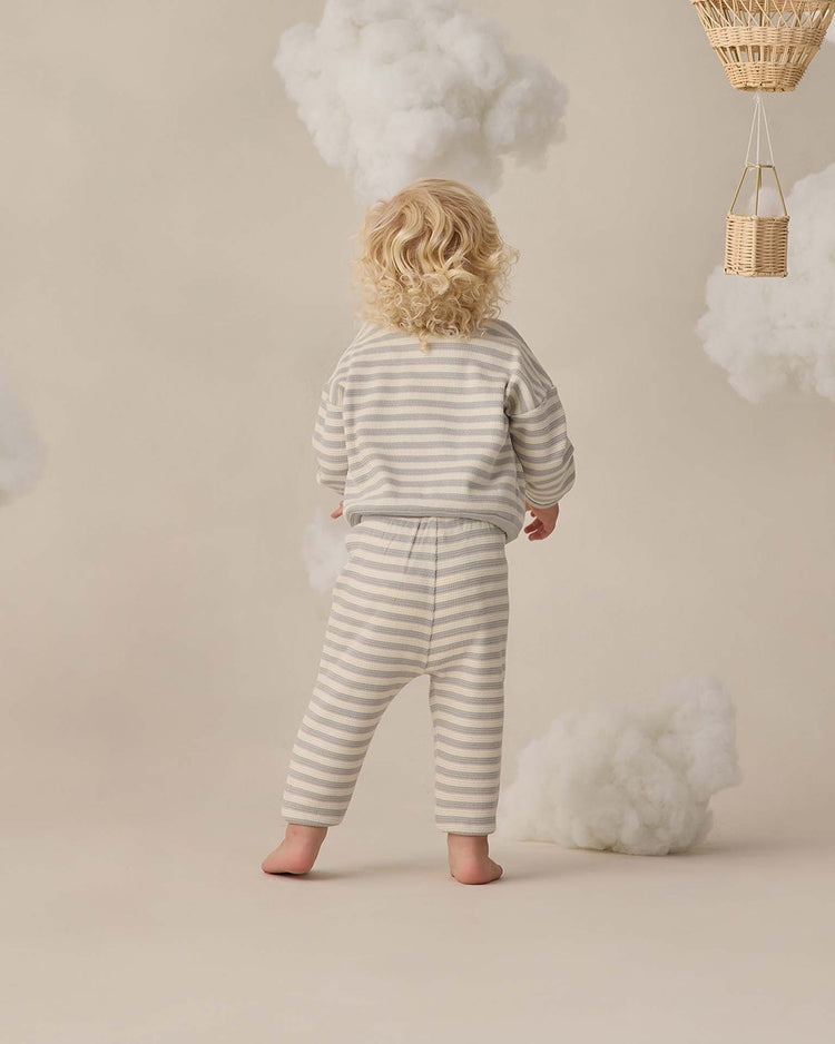 A young child with curly blonde hair stands barefoot, seen from behind, wearing the quincy mae waffle sweater set in sky stripe. Fluffy clouds and wicker baskets hang against a beige backdrop.