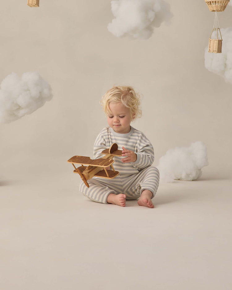 A toddler with blonde hair wears the quincy mae waffle sweater set in sky stripe while playing with a wooden airplane. Fluffy clouds and hot air balloons hang in the background, adding to the whimsical, dreamy scene.