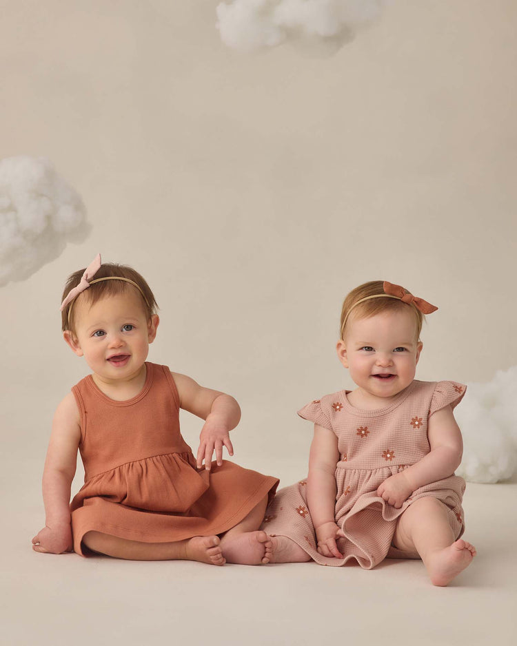 Two baby girls sit on the floor against a beige backdrop with fluffy clouds, both smiling. They wear matching headbands and quincy mae's ribbed tank dress in poppy—one plain, one floral—paired with cute bloomers.