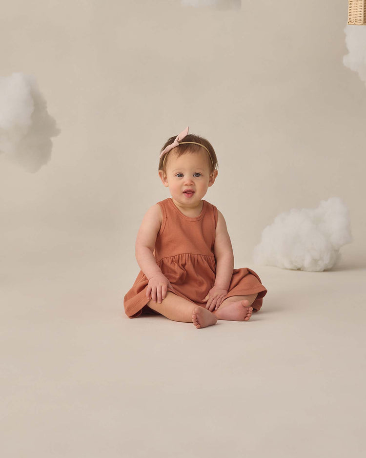 A baby wearing the quincy mae ribbed tank dress in poppy and a pink bow headband sits barefoot on a beige floor with fluffy white cloud props in the background.