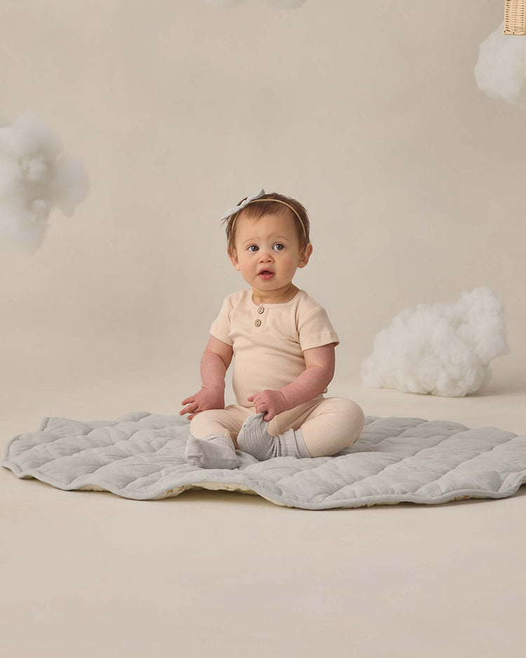 A baby in quincy mae’s ribbed legging in ecru sits on a soft light gray mat, surrounded by beige and fluffy cloud-like décor for a dreamy, whimsical vibe.