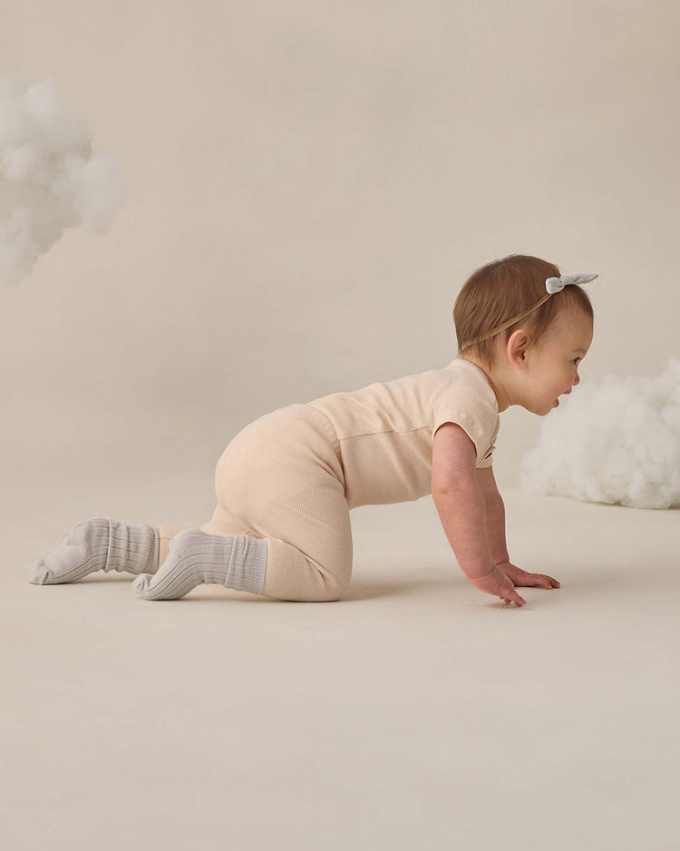 A baby in a beige onesie, gray socks, and a light headband crawls on a neutral floor with fluffy white clouds, wearing quincy mae's ribbed legging in ecru for extra comfort.