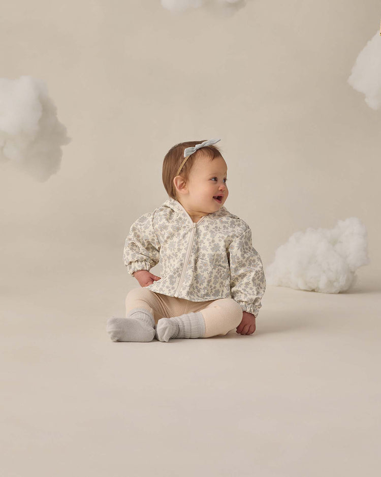 A baby in a floral jacket, quincy mae ribbed legging in ecru, socks, and a headband sits among cotton clouds on the floor, looking to the side and smiling against a beige background.