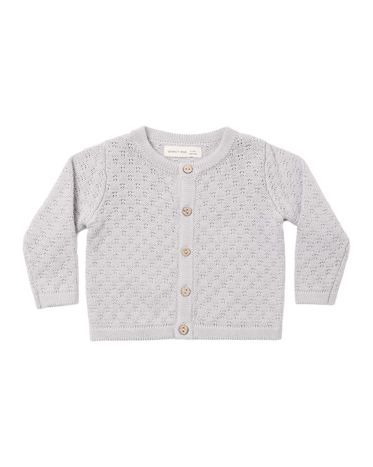 The knit cardigan in cloud by quincy mae is a light gray, long-sleeve baby cardigan made from organic cotton. It features five coconut buttons down the front, a delicate perforated textured knit pattern, and ribbed cuffs, hem, and neckline.
