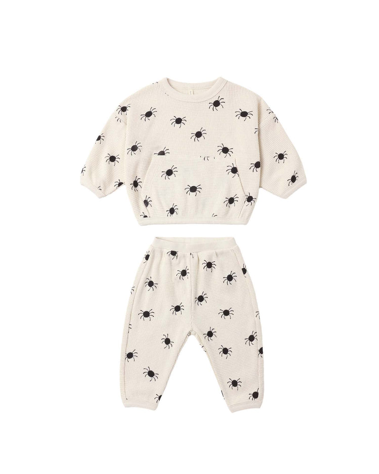 The waffle slouch set in bitsy spider by quincy mae is a cream-colored kids loungewear set, crafted from soft organic cotton. It features a short-sleeve top and matching pants with an all-over playful black bitsy spider print, shown laid flat on a white background—ideal for cozy lounging.