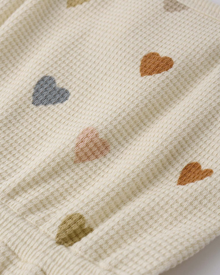 The quincy mae waffle cinch romper in confetti hearts features cream-colored, organic cotton waffle-knit fabric with small multicolored heart patterns—perfect for a cozy summer look.