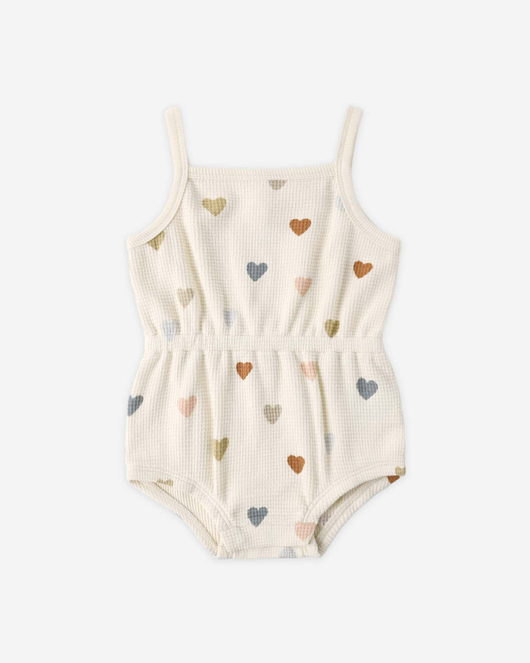 The quincy mae waffle cinch romper in confetti hearts features a sleeveless, cream design with thin straps, an elastic waist, and a pastel heart pattern, crafted from organic cotton—perfect for summer.