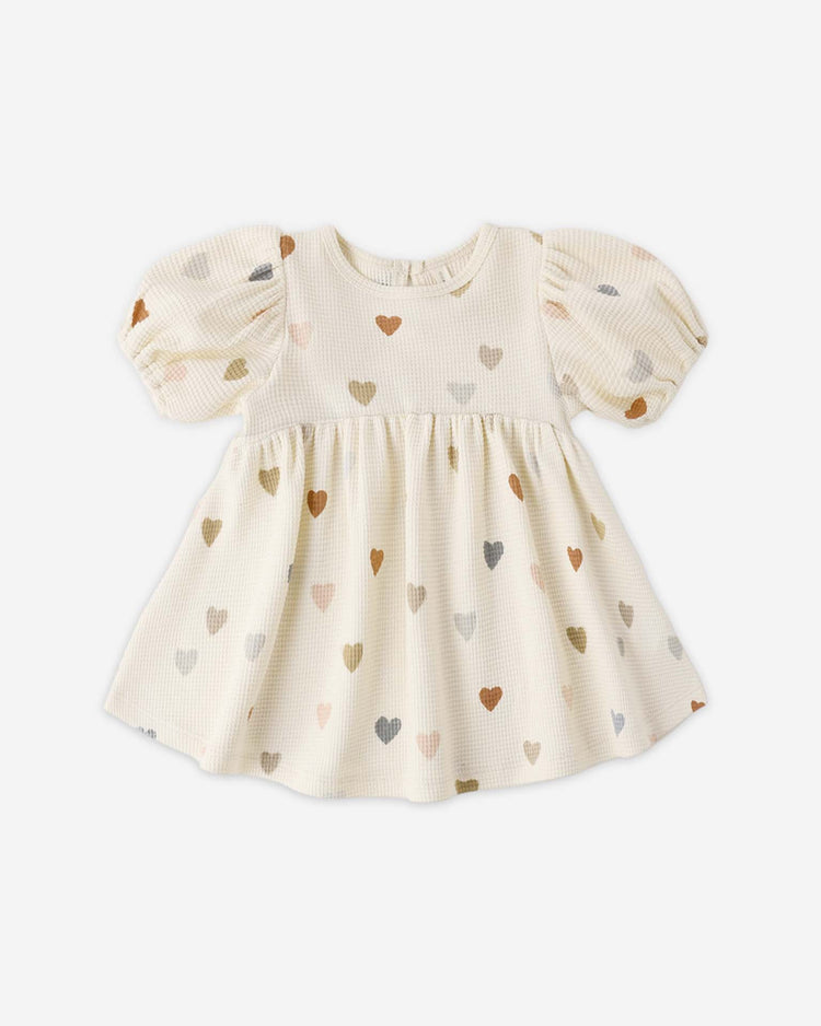 The quincy mae darla dress in confetti hearts is a cream-colored babydoll style with short puff sleeves, a flared skirt, and soft organic cotton. Adorned with tiny multicolored hearts, it's shown laid flat on a white background.