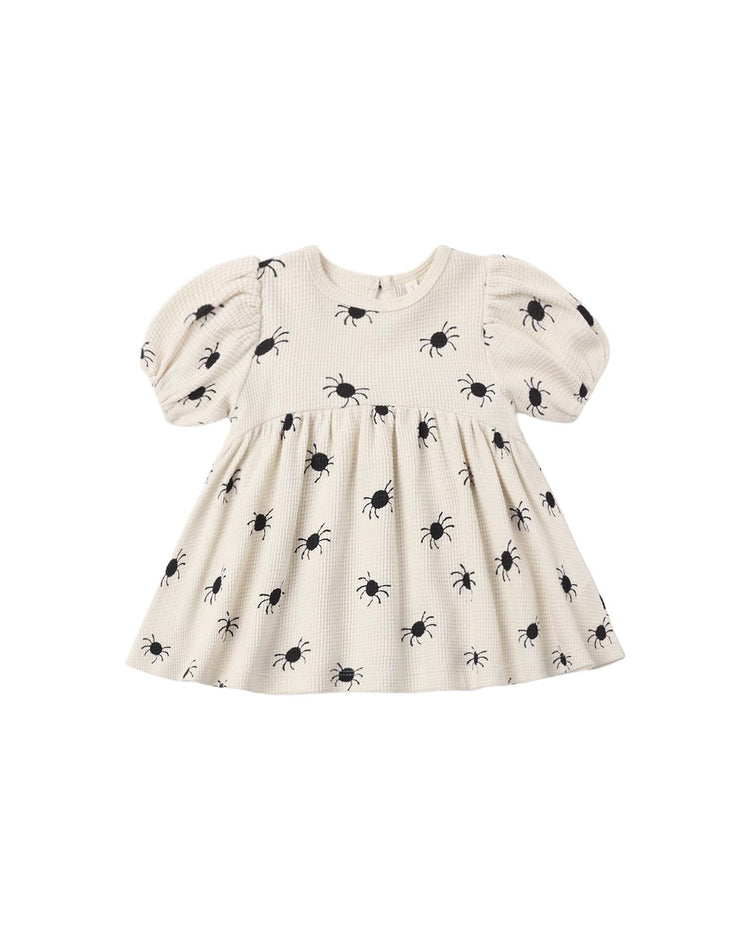 The quincy mae darla dress in bitsy spider is a cream-colored organic cotton baby dress featuring puffed short sleeves and an all-over pattern of evenly spaced black spiders.