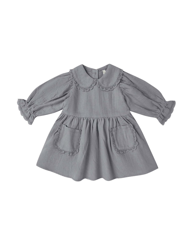The marisol dress in slate by quincy mae, crafted from soft organic cotton, features long sleeves, a Peter Pan collar, ruffled cuffs, and two front pockets with ruffled edges. The dress is displayed laid flat on a white background.