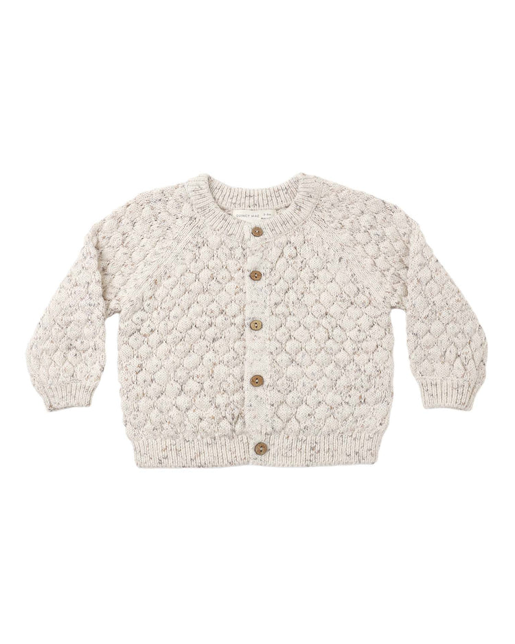 The bubble knit cardigan in natural confetti by quincy mae is a beige baby sweater crafted from soft organic cotton, showcasing a stylish diamond pattern with long sleeves, ribbed cuffs and hem, and five brown buttons down the front. This adorable cardigan is laid flat on a white background.