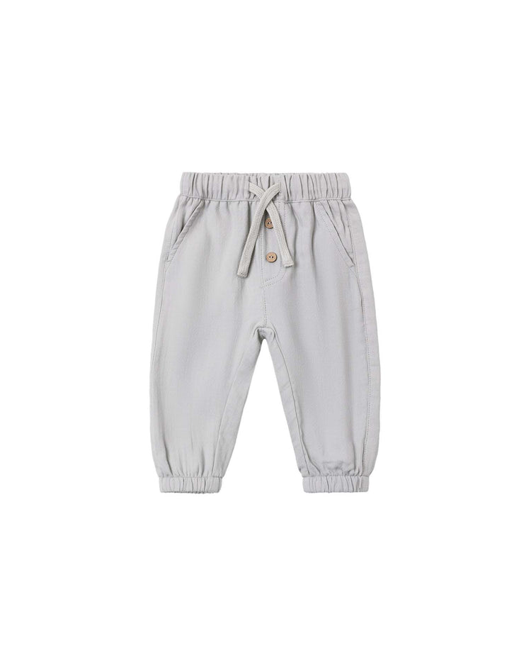 The giovani pant in cloud from quincy mae is crafted from soft organic cotton in a light gray woven gauze. These toddler pants feature an elastic waistband with a drawstring tie, two side pockets, a faux button fly, and elastic cuffs at the ankles. Shown laid flat on a white background.