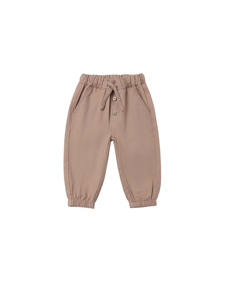 The giovani pant in almond by quincy mae is a pair of light brown toddler pants crafted from organic cotton, featuring an elastic waistband with a drawstring tie, two buttons, side pockets, and elastic cuffs at the ankles. The pants are shown laid flat on a white background.