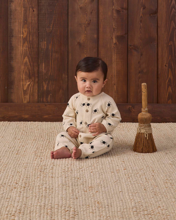 A baby sits on a beige textured rug, wearing the quincy mae waffle long sleeve jumpsuit in bitsy spider—an organic cotton outfit in cream with a playful spider print. A small broom stands upright beside the baby against a wooden wall backdrop.