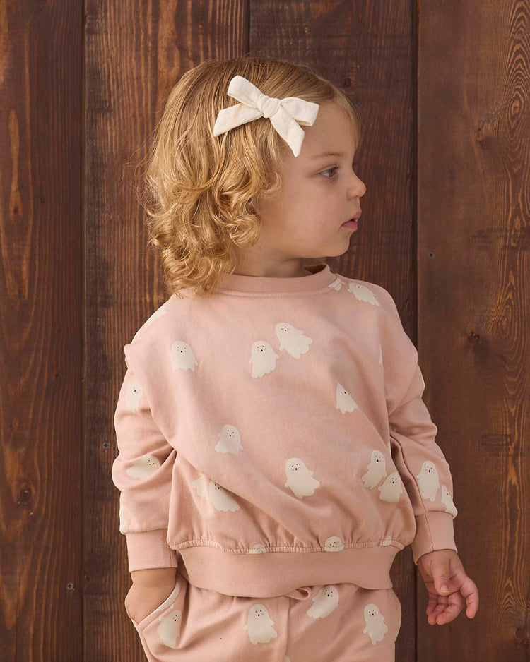 A young child with curly blond hair and a cream bow gazes to the side, wearing the Quincy Mae relaxed fleece sweatshirt in sweet ghost, standing in front of a wooden wall.