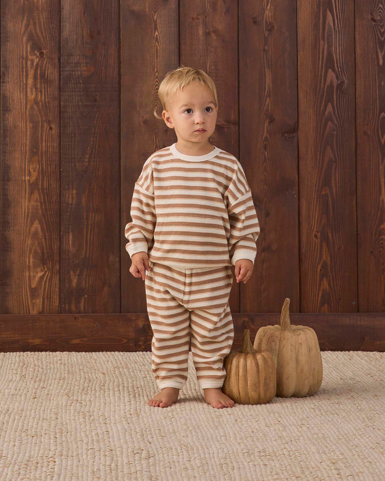 A toddler in the quincy mae waffle slouch set in spice stripe—organic cotton pajamas in beige and white stripes—stands barefoot on a textured rug beside two decorative pumpkins, with a wooden wall behind them.