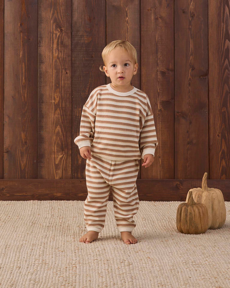 A toddler wearing the quincy mae waffle slouch set in spice stripe, crafted from organic cotton, stands barefoot on a textured rug beside two decorative wooden pumpkins, with a wooden wall as the background.