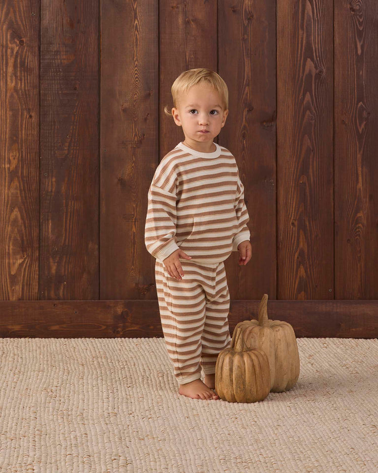 A young child wearing the quincy mae waffle slouch set in spice stripe, crafted from organic cotton, stands barefoot on a textured rug beside two decorative pumpkins, with a wooden wall as the backdrop.