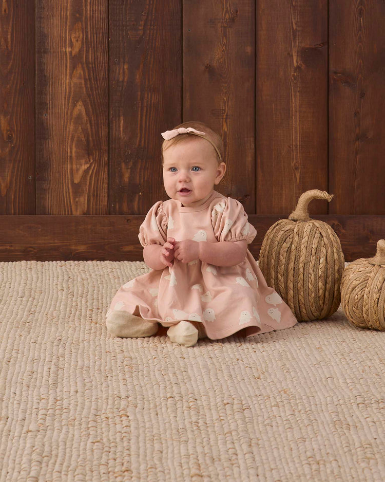 A baby in the quincy mae darla dress in spice stripe and matching headband sits on a textured rug next to two decorative, twine-wrapped pumpkins, with a wooden wall in the background.