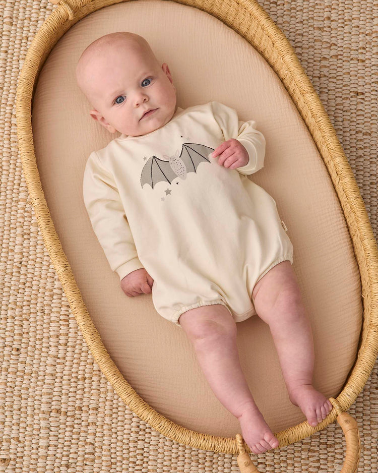 A baby in the quincy mae crewneck bubble romper in baby bat—an organic cotton, cream-colored outfit featuring a playful bat graphic—lies in a woven Moses basket atop a textured beige mat, gazing upward with wide blue eyes.
