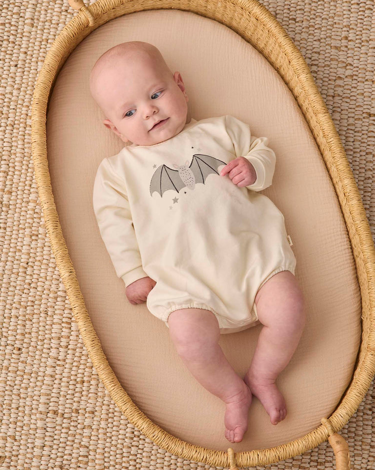 A baby wearing the quincy mae crewneck bubble romper in baby bat lies on a beige oval basket atop a textured woven mat, looking slightly upward.