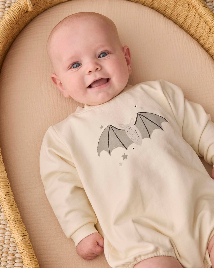 A smiling baby lies in a woven basket, wearing the quincy mae crewneck bubble romper in baby bat—an organic cotton, cream-colored outfit featuring an adorable bat and stars design on the chest.