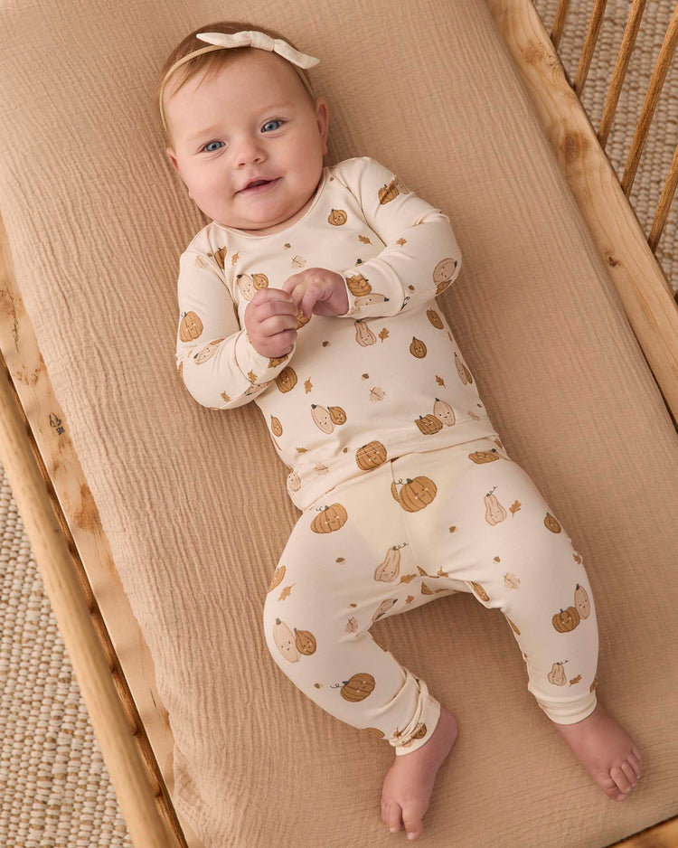 A baby smiles and looks up while lying on a beige blanket in a crib, dressed in the Quincy Mae bamboo pajama set in harvest friends with a matching bow headband.