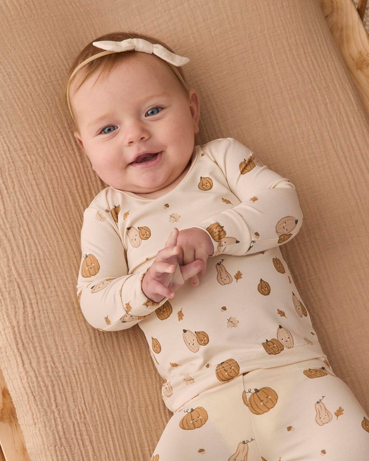 A baby with a light bow headband smiles while lying on a beige blanket, wearing the quincy mae bamboo pajama set in harvest friends.