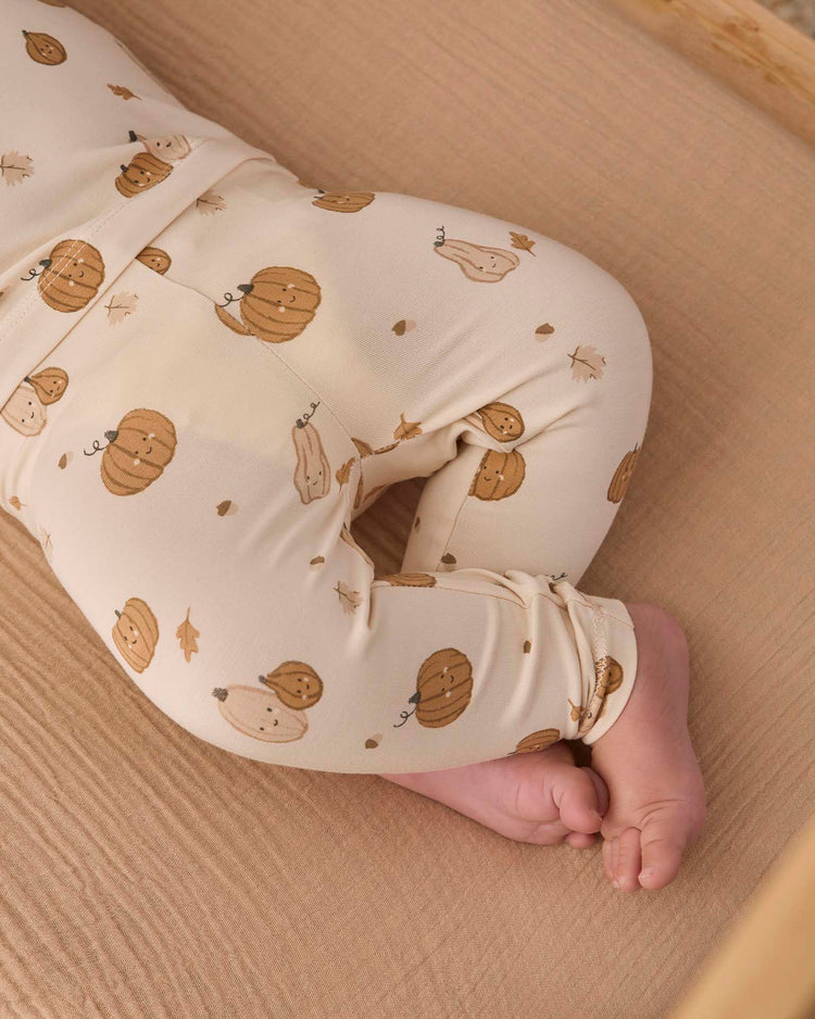 A baby lies on a beige sheet, wearing the Quincy Mae bamboo pajama set in Harvest Friends—a cream-colored slim-fit style featuring brown pumpkin and leaf patterns. Only the lower half of the baby’s body and feet are visible.