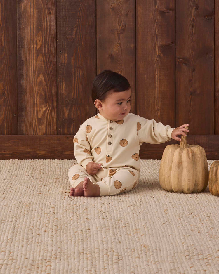 A baby in the quincy mae relaxed fleece jumpsuit in pumpkins, made from cozy organic cotton, sits on a textured rug and reaches for a light-colored pumpkin, with a wooden wall behind them.