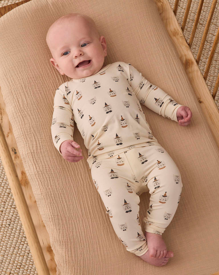 A baby in a Quincy Mae bamboo pajama set in Costume Kittens, featuring a slim fit and long sleeves, lies smiling on a beige padded mat on a wooden crib.