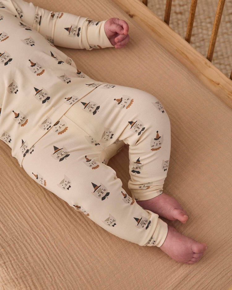A baby wearing the quincy mae bamboo pajama set in costume kittens, featuring a slim-fit long sleeve design with playful kitten faces, lies on a tan crib sheet with the wooden slats of the crib visible. The baby’s face is not shown—only the legs and lower body are visible.