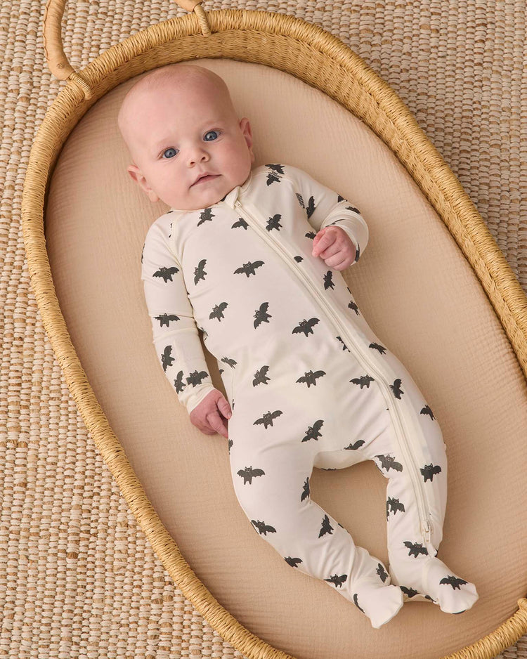 A baby in the quincy mae bamboo zip footie in bats lies in a woven bassinet on a textured rug, looking up with wide blue eyes, cozy in their soft outfit adorned with black bat prints.