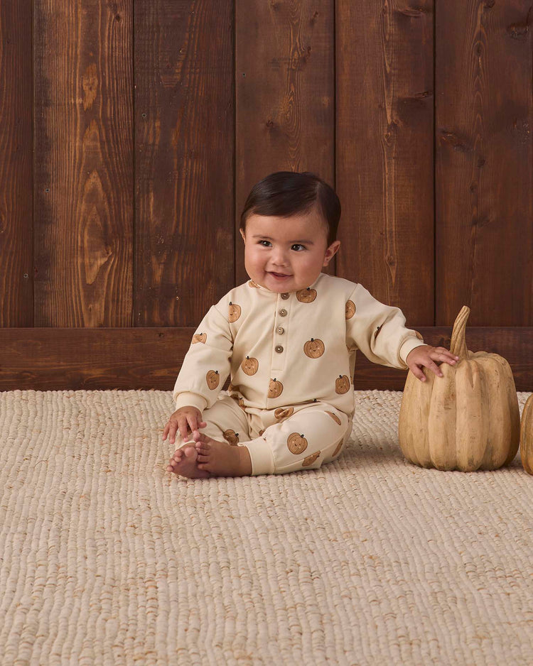A baby wearing the quincy mae relaxed fleece jumpsuit in pumpkins sits on a woven rug, smiling, with one hand resting on a decorative pumpkin. A wooden wall and another pumpkin are visible in the background.