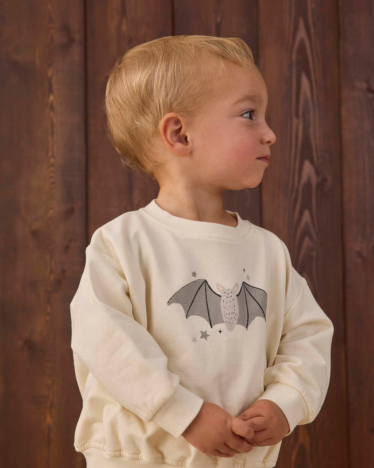 A young child with blond hair stands in front of a wooden wall, wearing the quincy mae relaxed fleece sweatshirt in baby bats—a cream-colored organic cotton top featuring a baby bat and stars graphic on the front. The child is looking to the right with hands clasped together.