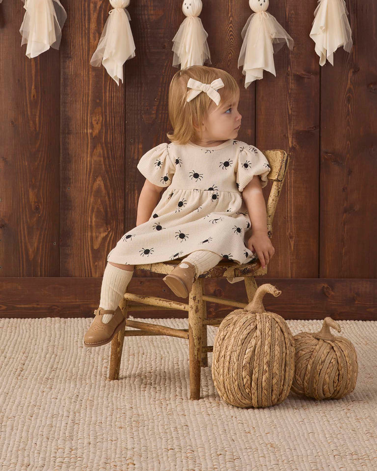 A toddler wearing the quincy mae darla dress in bitsy spider, made from cream organic cotton with black prints, sits on a rustic wooden chair and gazes off to the side. Two woven pumpkins rest on the floor nearby, while ghost decorations hang from a wooden wall in the background.