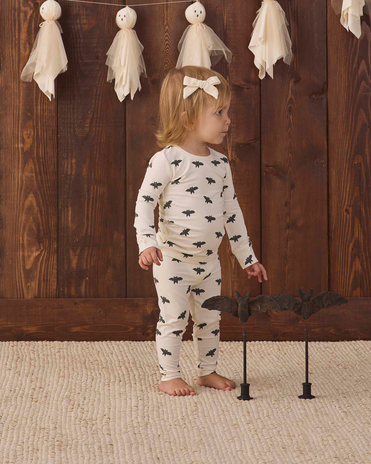 A toddler wearing the quincy mae bamboo pajama set in bats stands on a textured rug, gazing to the side. Behind her, a wooden wall is decorated with ghost ornaments, and two bat props on stands are positioned in front of her.