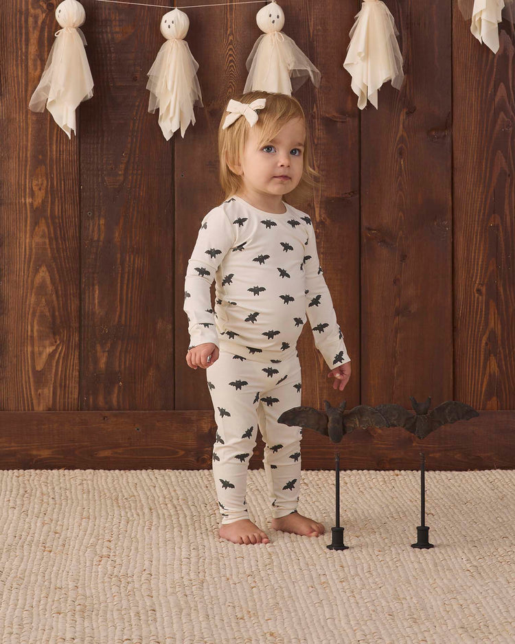 A toddler wearing the quincy mae bamboo pajama set in bats stands on a textured rug with a bat decoration nearby, in front of a wooden wall adorned with white ghost garlands.