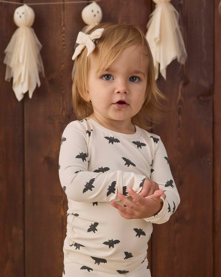 A toddler with light brown hair and a bow wears the quincy mae bamboo pajama set in bats, a slim-fit, cute bat-printed outfit. She stands in front of a wooden wall decorated with ghost-shaped garlands.